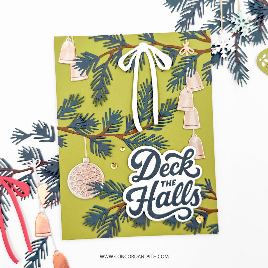 A handmade Christmas greeting card with a green background, layered pine branches, hanging beige bells, and the phrase 'Deck the Halls' in layered white and navy lettering. The card is styled on a white background with holiday paper decorations around it.