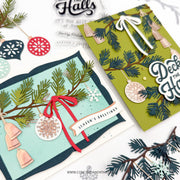 Two handmade layered Christmas greeting cards displayed on a white background with festive crafting props. One card is light aqua with green pine branches, copper bells, a red ribbon, and a white banner reading 'Season's Greetings.