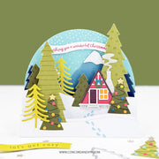 A 3D layered pop-up Christmas card featuring a cozy cabin surrounded by colorful pine trees under a blue snowy sky. The card is displayed on a white surface against a muted olive green background.