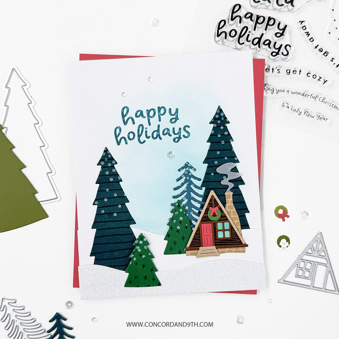 A handmade layered holiday card featuring a small cabin, pine trees, and the words 'happy holidays' in blue script. The card is styled on a white background with craft dies, stamps, and other cards around it.