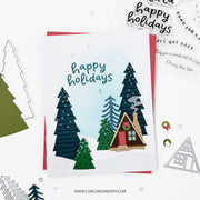 A handmade layered holiday card featuring a small cabin, pine trees, and the words 'happy holidays' in blue script. The card is styled on a white background with craft dies, stamps, and other cards around it.
