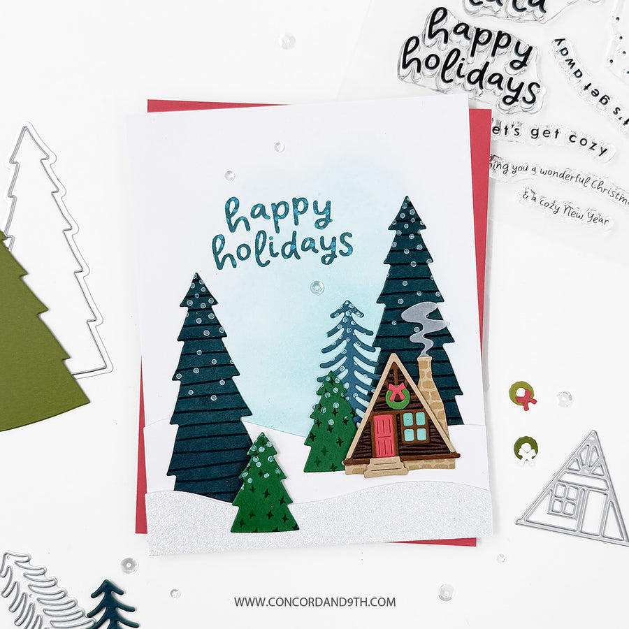 A handmade layered holiday card featuring a small cabin, pine trees, and the words 'happy holidays' in blue script. The card is styled on a white background with craft dies, stamps, and other cards around it.