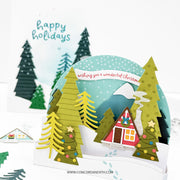 A handmade pop-up cardboard Christmas card featuring a snowy winter village scene with layered green and yellow pine trees, a red cabin, mountains, and a banner reading 'wishing you a wonderful Christmas.' A second card in the background says 'happy holidays.