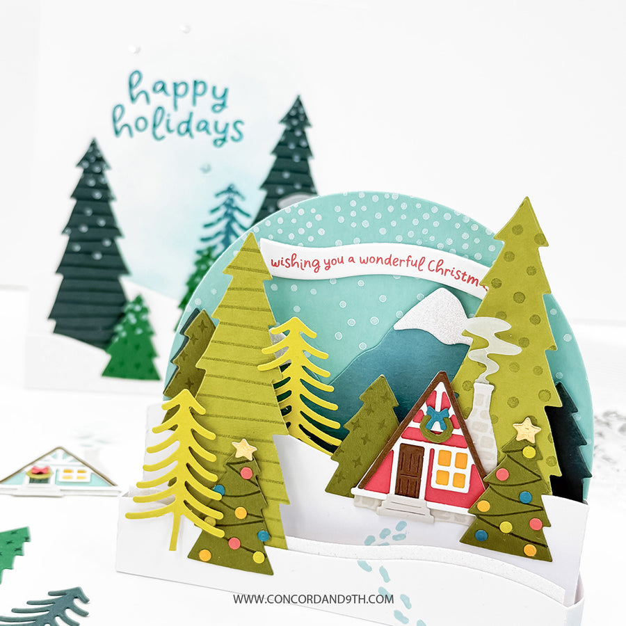 A handmade pop-up cardboard Christmas card featuring a snowy winter village scene with layered green and yellow pine trees, a red cabin, mountains, and a banner reading 'wishing you a wonderful Christmas.' A second card in the background says 'happy holidays.
