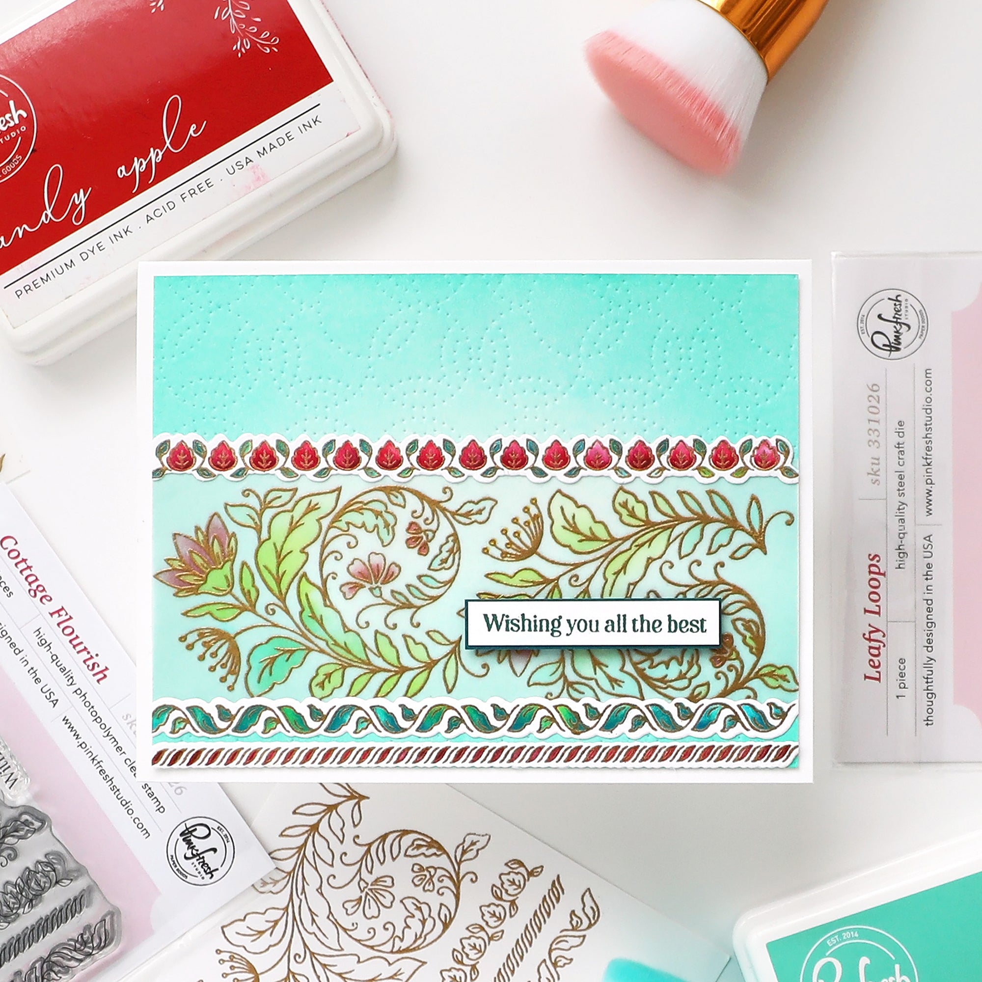 A handmade layered card with floral and leaf patterns in mint green, gold, and red, featuring a central white banner that reads 'Wishing you all the best'. The card is styled in a flat-lay arrangement with crafting supplies including an 'Apple Pie' die, a 'Cottage Flavour' ink pad, and a 'Leaf Loops' stamp card.