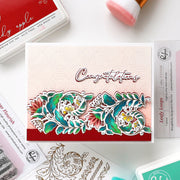 A handmade congratulations card featuring a layered floral die-cut design in teal, green, pink, and yellow with the word 'Congratulations' in white script. The card is styled in a flat-lay arrangement with crafting supplies including an Appleton ink pad, a pink brush, and a Leafy Loops stamp package.