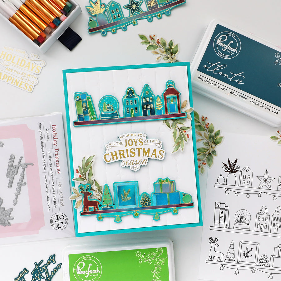A flat-lay of a handmade Christmas-themed card featuring layered die-cut elements in teal, blue, green, and gold, surrounded by craft supplies including an ink pad, stamps, and coloring pencils on a white background.