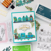 A flat-lay of a handmade Christmas-themed card featuring layered die-cut elements in teal, blue, green, and gold, surrounded by craft supplies including an ink pad, stamps, and coloring pencils on a white background.