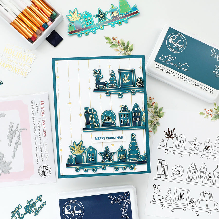 A styled flat-lay of crafting supplies on a white surface, featuring a handmade Christmas card with teal layered die-cut elements, an ink pad, stamp sheets, colored pencils, and small decorative botanicals. The card and packaging show the brand name Regfachs.