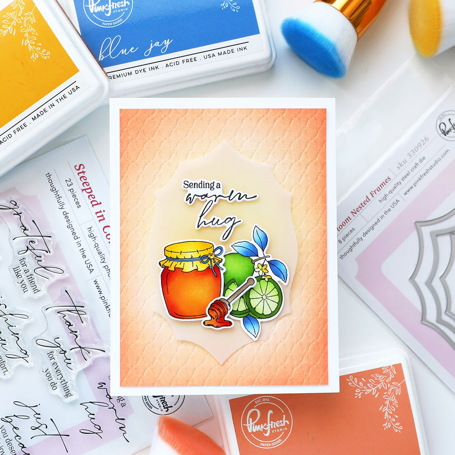 A handmade greeting card featuring a honey jar, lime slices, and a blue flower with the text 'Sending a warm hug' on a peach textured background, surrounded by crafting supplies including Pouchfresh ink pads and a makeup brush.