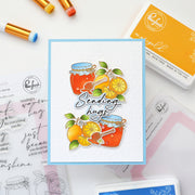 A handmade greeting card featuring illustrated honey jars, oranges, and the phrase 'Sending hugs' in script, styled on a white surface with craft supplies including stamps, ink pads, and paint pens.