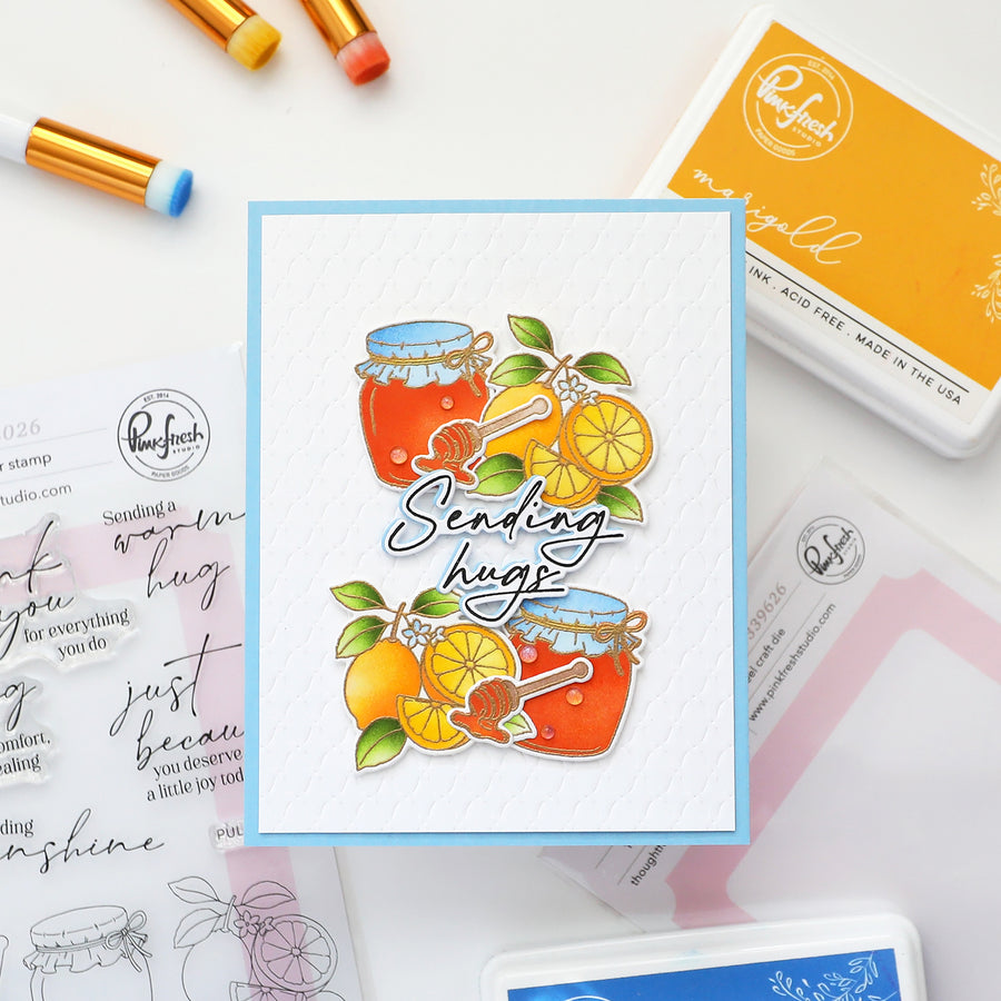 A handmade greeting card featuring illustrated honey jars, oranges, and the phrase 'Sending hugs' in script, styled on a white surface with craft supplies including stamps, ink pads, and paint pens.