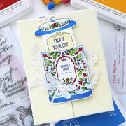 A handmade greeting card featuring a whimsical illustrated bottle with the words 'ENJOY YOUR DAY' and 'SIMPLE JOYS,' layered with white floral die-cut elements on a pale cream cardstock. The card is photographed on a desk with craft supplies visible around it, including markers, a stamp sheet, and printed cards.