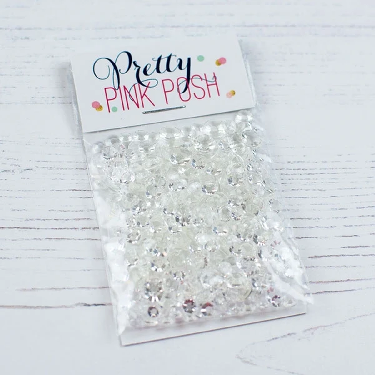 A clear plastic bag filled with many small clear rhinestones or crystal embellishments, attached to a white paper header card that reads 'Pretty PINK POH' in dark script and pink block letters. The package is photographed on a white wood surface. Pretty Pink Posh Sparkling Clear Jewels