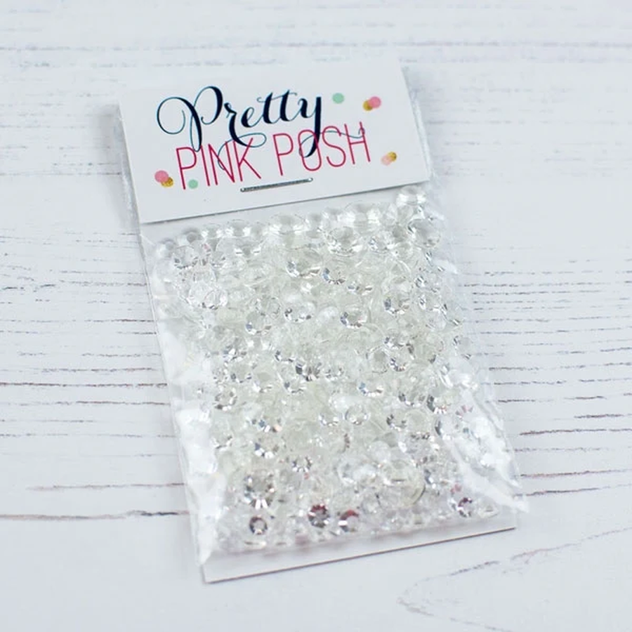 A clear plastic bag filled with many small clear rhinestones or crystal embellishments, attached to a white paper header card that reads 'Pretty PINK POH' in dark script and pink block letters. The package is photographed on a white wood surface. Pretty Pink Posh Sparkling Clear Jewels