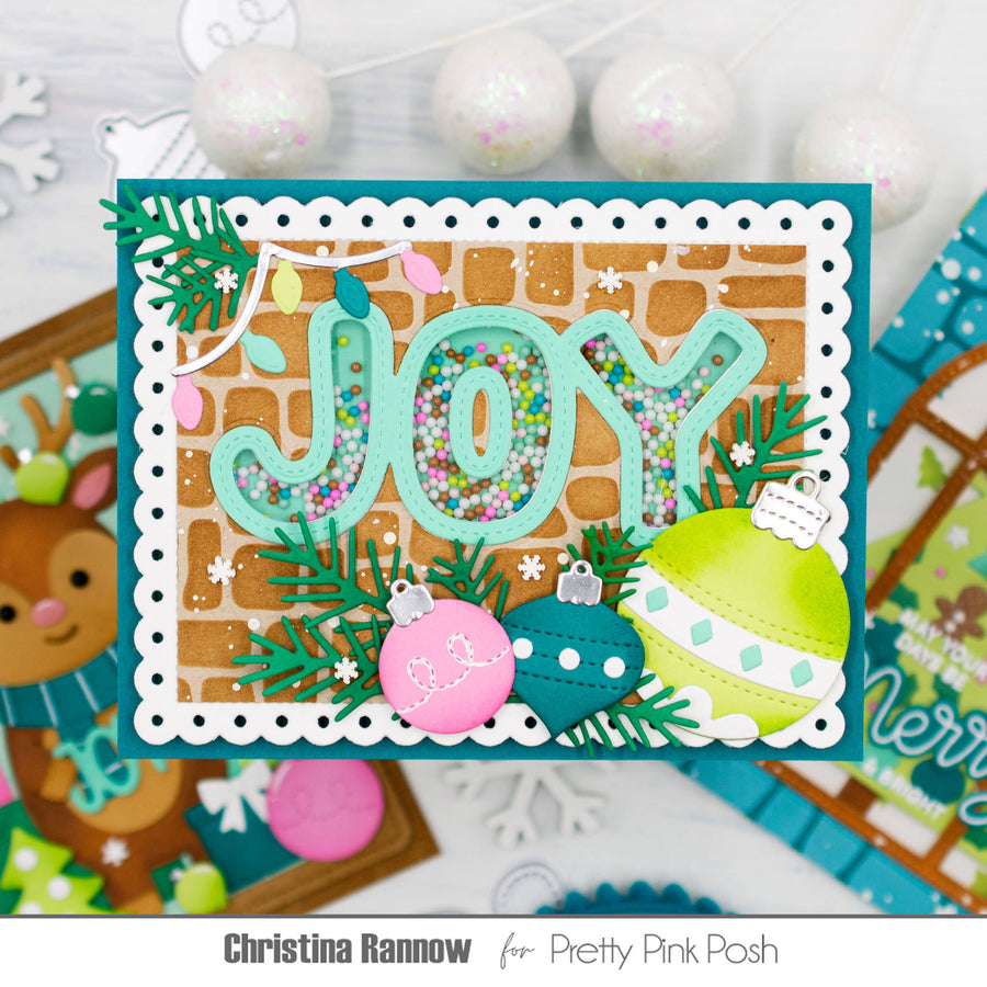 A handmade Christmas greeting card featuring the word 'JOY' in layered mint green letters filled with multicolored confetti, decorated with green pine branches, a pink ornament, a lime green ornament, and a white scalloped border. The card is displayed on a white surface with holiday-themed props in the background, and text at the bottom reads 'Christina Rannow for Pretty Pink Posh'.