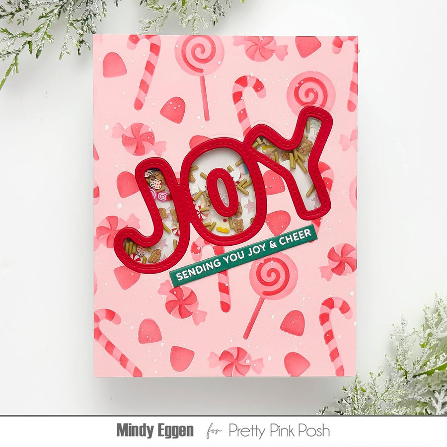 A handmade pink holiday greeting card featuring the word 'JOY' in 3D red letters with a window showing gold glitter and small illustrations. The card has a candy cane and lollipop pattern background and a green banner that reads 'SENDING YOU JOY & CHEER'.