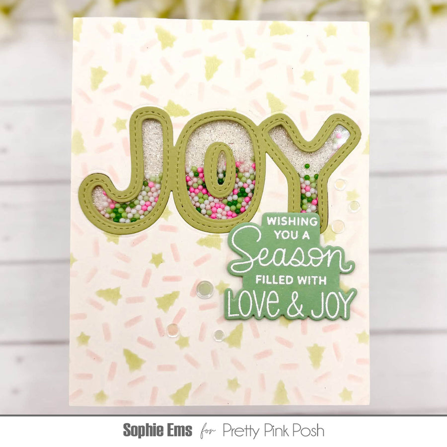A handmade greeting card featuring the word 'JOY' in large olive-green letters filled with glitter and small pink, green, and white beads. The card has a white background with light pink and yellow star confetti, and a layered green badge that reads 'WISHING YOU A Season FILLED WITH LOVE & JOY'.