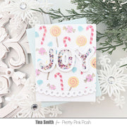 A handmade greeting card featuring the word 'JOY' in glittery letters, decorated with candy canes, lollipops, and pastel shapes, styled on a light background with white ornaments and evergreen branches.