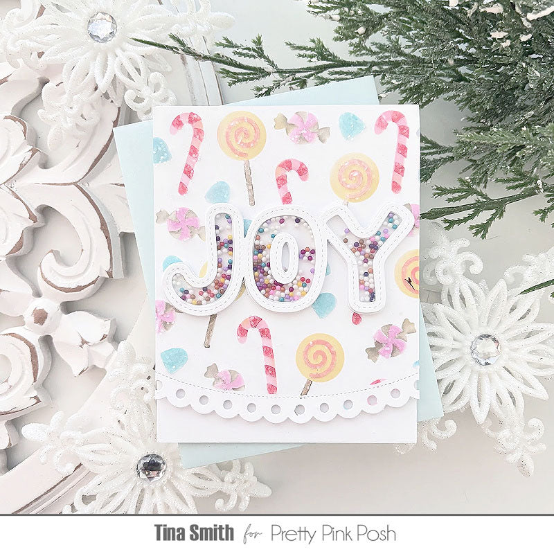 A handmade greeting card featuring the word 'JOY' in glittery letters, decorated with candy canes, lollipops, and pastel shapes, styled on a light background with white ornaments and evergreen branches.