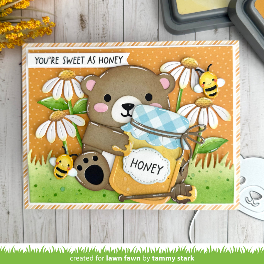 A handmade greeting card featuring a cute bear holding a jar of honey, surrounded by daisies, bees, and the text 'YOU'RE SWEET AS HONEY'. The card is displayed on a light gray wooden surface with crafting tools partially visible in the corners. Lawn Fawn Big Hugs Pop-up Dies