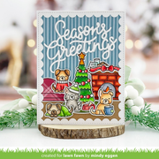 A handmade holiday greeting card featuring cartoon dogs around a Christmas tree and a fireplace, with the text 'Seasons Greetings' on a blue striped background. The card is displayed on a wood slice coaster with blurred holiday greenery and lights in the background.