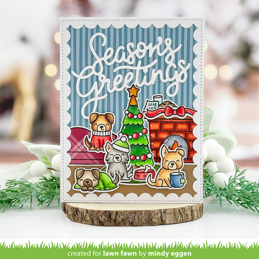 A handmade holiday greeting card featuring cartoon dogs around a Christmas tree and a fireplace, with the text 'Seasons Greetings' on a blue striped background. The card is displayed on a wood slice coaster with blurred holiday greenery and lights in the background.