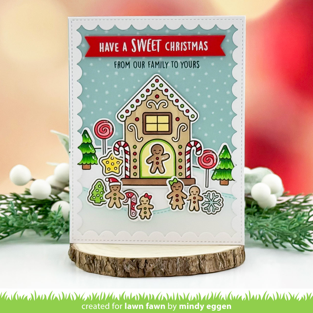 A handmade Christmas greeting card featuring a gingerbread house design with candy canes, lollipops, pine trees, and the text 'HAVE A SWEET CHRISTMAS FROM OUR FAMILY TO YOURS'. The card is displayed on a rustic wood slice stand against a softly blurred holiday-themed background with greenery and white berries.