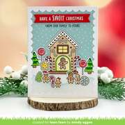 A handmade Christmas greeting card featuring a gingerbread house design with candy canes, lollipops, pine trees, and the text 'HAVE A SWEET CHRISTMAS FROM OUR FAMILY TO YOURS'. The card is displayed on a rustic wood slice stand against a softly blurred holiday-themed background with greenery and white berries.