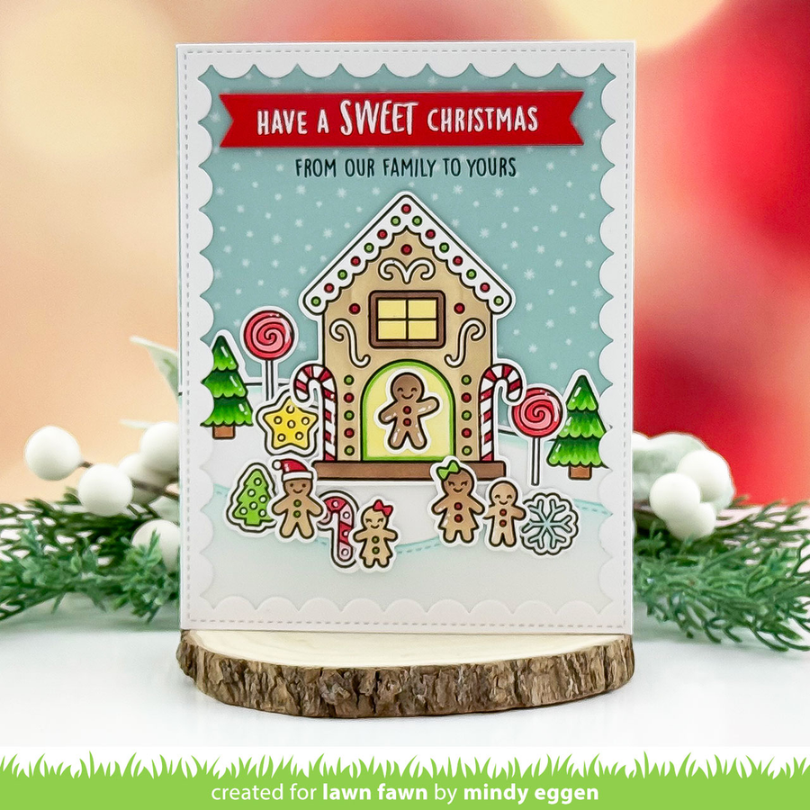 A handmade Christmas greeting card featuring a gingerbread house design with candy canes, lollipops, pine trees, and the text 'HAVE A SWEET CHRISTMAS FROM OUR FAMILY TO YOURS'. The card is displayed on a rustic wood slice stand against a softly blurred holiday-themed background with greenery and white berries.