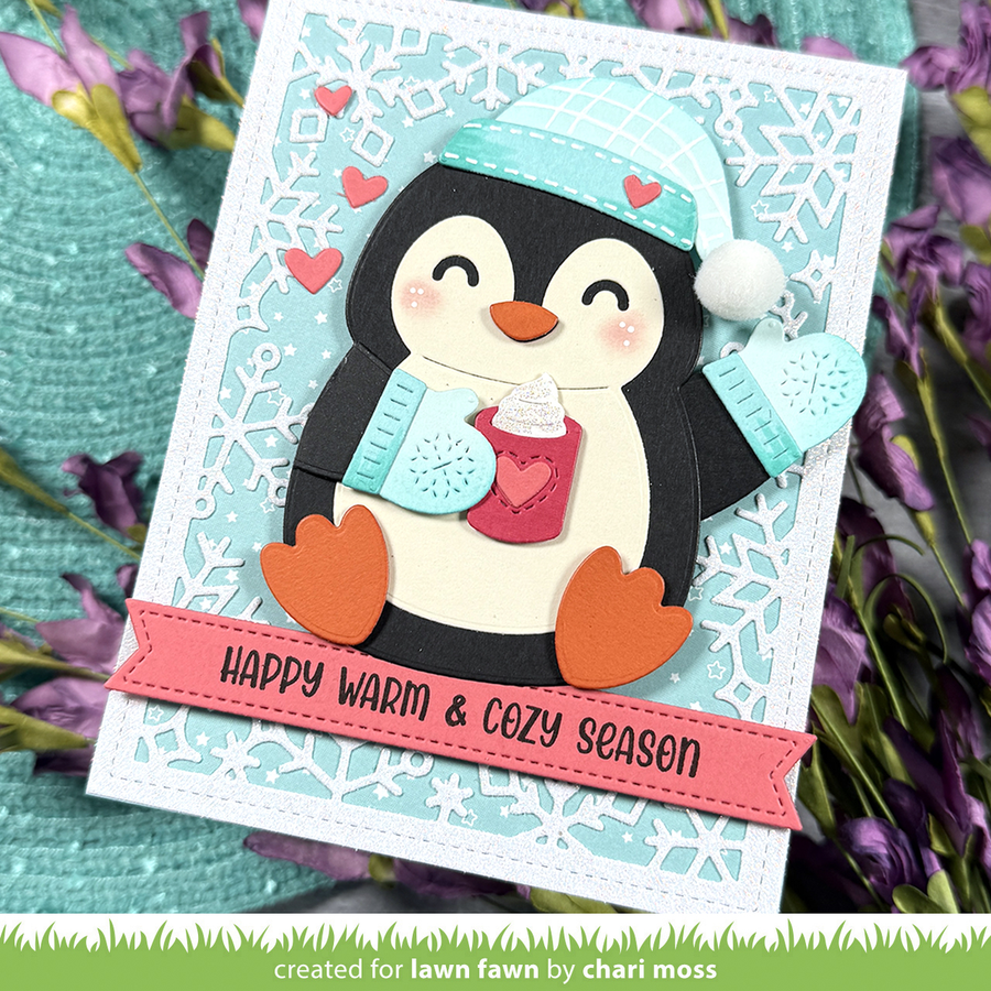 A handmade penguin card featuring a layered paper penguin wearing a light blue plaid hat and matching mittens, holding a red mug with whipped cream. The card has a white scalloped border, pink hearts, and a pink banner reading 'HAPPY WARM & COZY SEASON'.