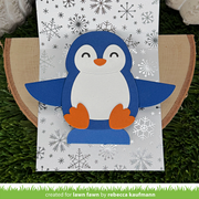 A handmade penguin card featuring a blue, white, and orange paper cutout penguin sitting on a snowy white card with silver snowflake patterns, displayed on a wood slice over artificial green grass.