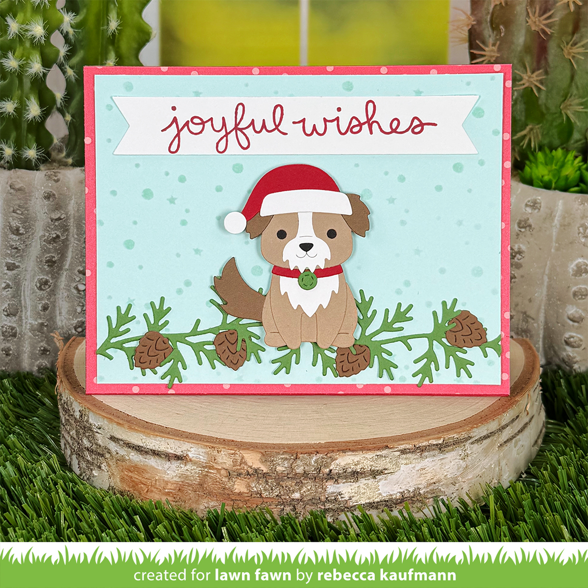 A handmade Christmas greeting card featuring a cute brown dog wearing a red Santa hat, set against a light blue background with pine branches and the words 'joyful wishes' in red script. The card is displayed on a rustic birch log slice with artificial grass and cacti in the background.