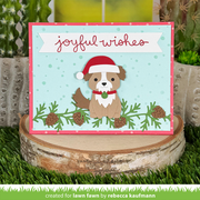 A handmade Christmas greeting card featuring a cute brown dog wearing a red Santa hat, set against a light blue background with pine branches and the words 'joyful wishes' in red script. The card is displayed on a rustic birch log slice with artificial grass and cacti in the background.