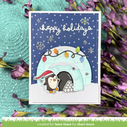 A handmade holiday greeting card featuring a penguin in a Santa hat next to a light blue arched igloo with colorful string lights and snowflakes on a dark blue background. The card is displayed on a textured mint green surface with purple flowers in the background.