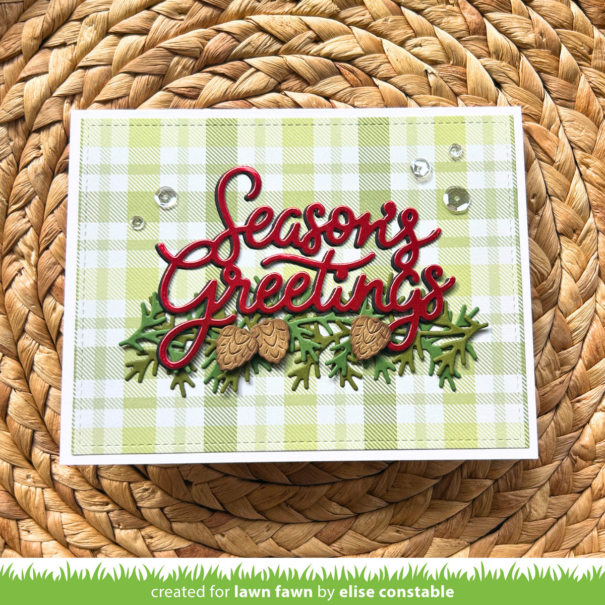 A handmade greeting card with a light green and white gingham pattern, featuring red cursive 'Seasons Greetings' text, green pine branches, and two pinecones, displayed on a woven straw mat.