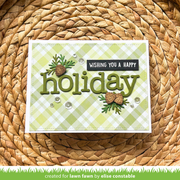 A handmade greeting card with a light green and white gingham pattern, featuring the word 'holiday' in olive green letters, pinecones, greenery, and a black banner that reads 'WISHING YOU A HAPPY holiday'. The card is placed on a round woven straw mat, with a green banner at the bottom reading 'created for lawn fawn by elise constable'.