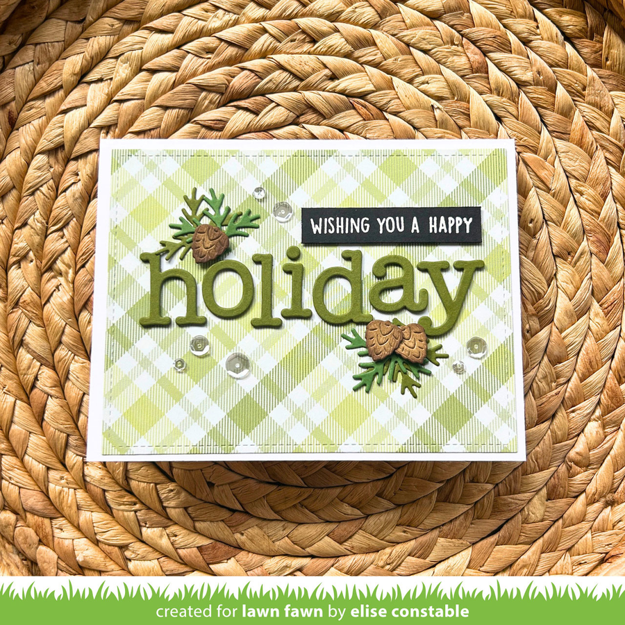 A handmade greeting card with a light green and white gingham pattern, featuring the word 'holiday' in olive green letters, pinecones, greenery, and a black banner that reads 'WISHING YOU A HAPPY holiday'. The card is placed on a round woven straw mat, with a green banner at the bottom reading 'created for lawn fawn by elise constable'.