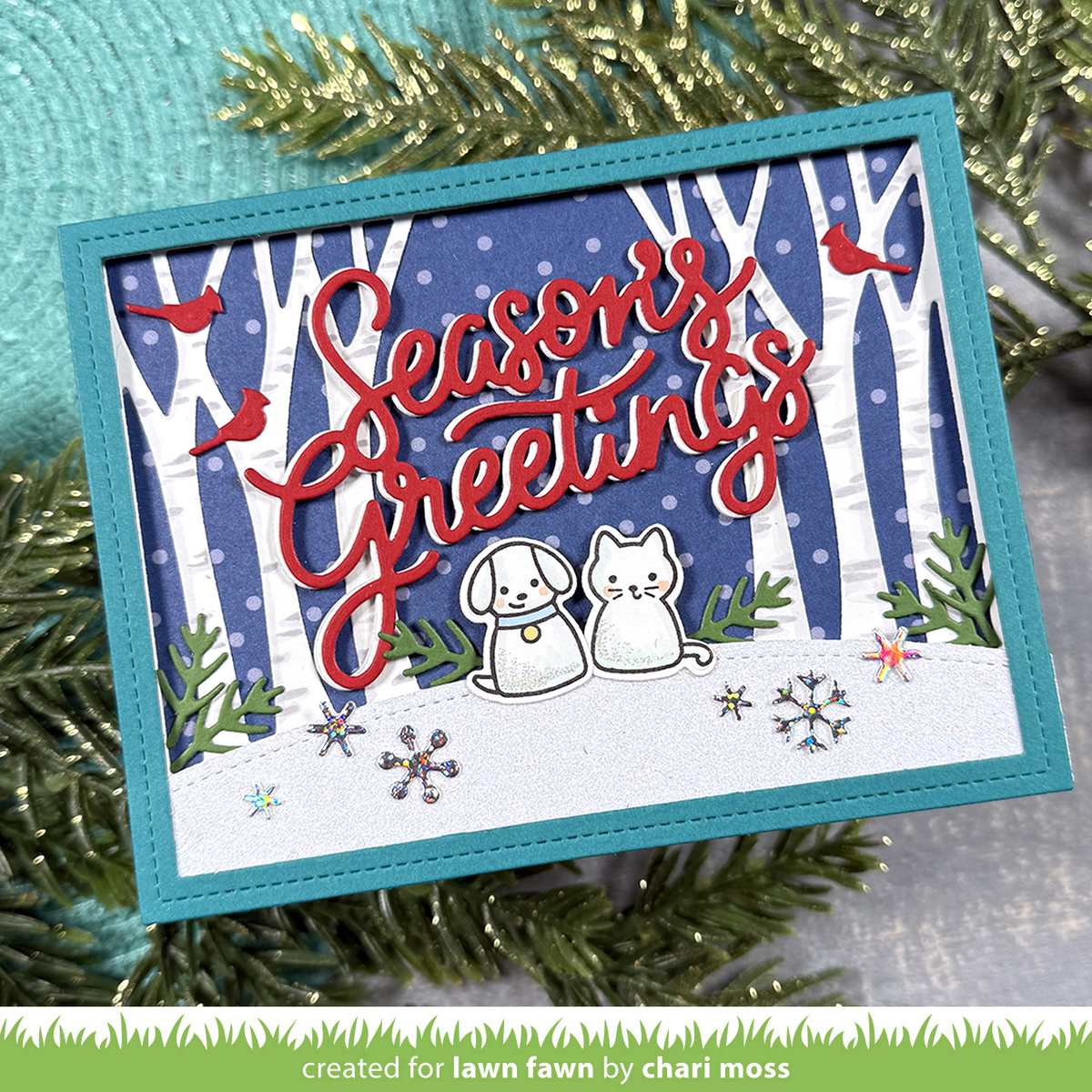 A handmade layered greeting card with a turquoise border, featuring a winter forest scene with white birch trees, red birds, a blue polka-dot background, a white snow-covered ground, and red script text reading 'Seasons Greetings.' Two small die-cut animal figures sit in the snow, surrounded by greenery and snowflake embellishments.