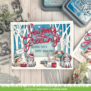 A handmade layered card featuring cute cartoon dogs and cats in winter clothing, with red cursive text reading 'Season's Greetings' and a white message bubble that says 'Wishing you a Happy Howliday.' The card is styled on a whitewashed wood surface with stamp dies, ink pads, and small holiday greenery around it.