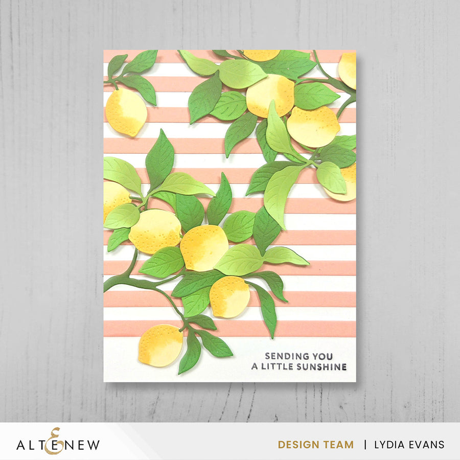 A rectangular greeting card or art print featuring yellow lemons and green leaves over soft peach-and-white horizontal stripes, with the text 'SENDING YOU A LITTLE SUNSHINE' at the bottom. The card is photographed against a light gray wood-textured background, with ALTENEW branding and 'Design Team | Lydia Evans' visible at the bottom of the image. Altenew Creative Cuts: Lemon Tree Layering Die Set card ideas