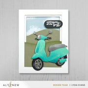 A framed handmade greeting card featuring a turquoise scooter layered over olive green hills under a light blue sky, with a black speech bubble that reads 'wishing you all the best.' The card is displayed against a light gray wood-textured background, with the brand name ALTENEW and designer name Lydia Evans visible at the bottom. Altenew Nesting A2 Panels Die Set card ideas