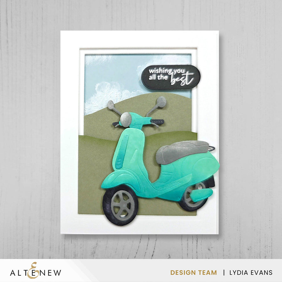 A framed handmade greeting card featuring a turquoise scooter layered over olive green hills under a light blue sky, with a black speech bubble that reads 'wishing you all the best.' The card is displayed against a light gray wood-textured background, with the brand name ALTENEW and designer name Lydia Evans visible at the bottom. Altenew Nesting A2 Panels Die Set card ideas