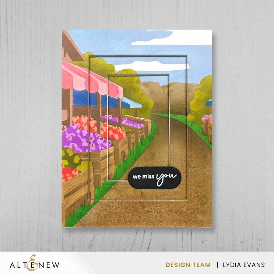 A handmade greeting card or art print featuring a stylized illustrated farm market scene with flower boxes, a dirt path, trees, and a blue sky. The card has layered geometric frame lines and a black label reading 'we miss you'. Altenew Nesting A2 Panels Die Set card ideas