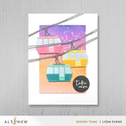 A handmade greeting card featuring colorful cable cars on a purple-to-orange gradient background, displayed on a light gray wood surface. The card has a white border and a black circular sticker that reads 'take care'. Altenew One-Go: Solid Circular Sentiments Press Plate card ideas