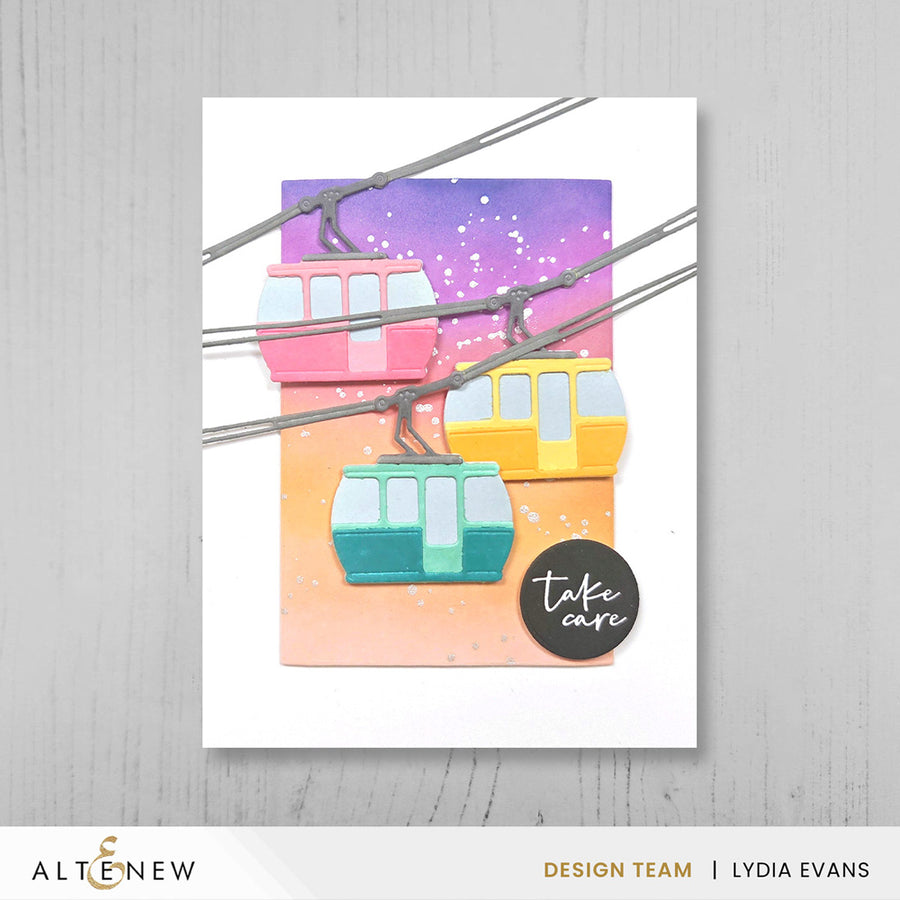 A handmade greeting card featuring colorful cable cars on a purple-to-orange gradient background, displayed on a light gray wood surface. The card has a white border and a black circular sticker that reads 'take care'. Altenew One-Go: Solid Circular Sentiments Press Plate card ideas