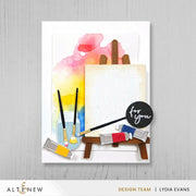 A handmade greeting card featuring a watercolor painting palette design with paintbrushes, paint tubes, a blank canvas on an easel, and a black circular badge that reads 'for you'. The card is displayed against a light gray wood-textured background, with the Altenew and Design Team Lydia Evans branding visible. Altenew One-Go: Solid Circular Sentiments Press Plate card ideas