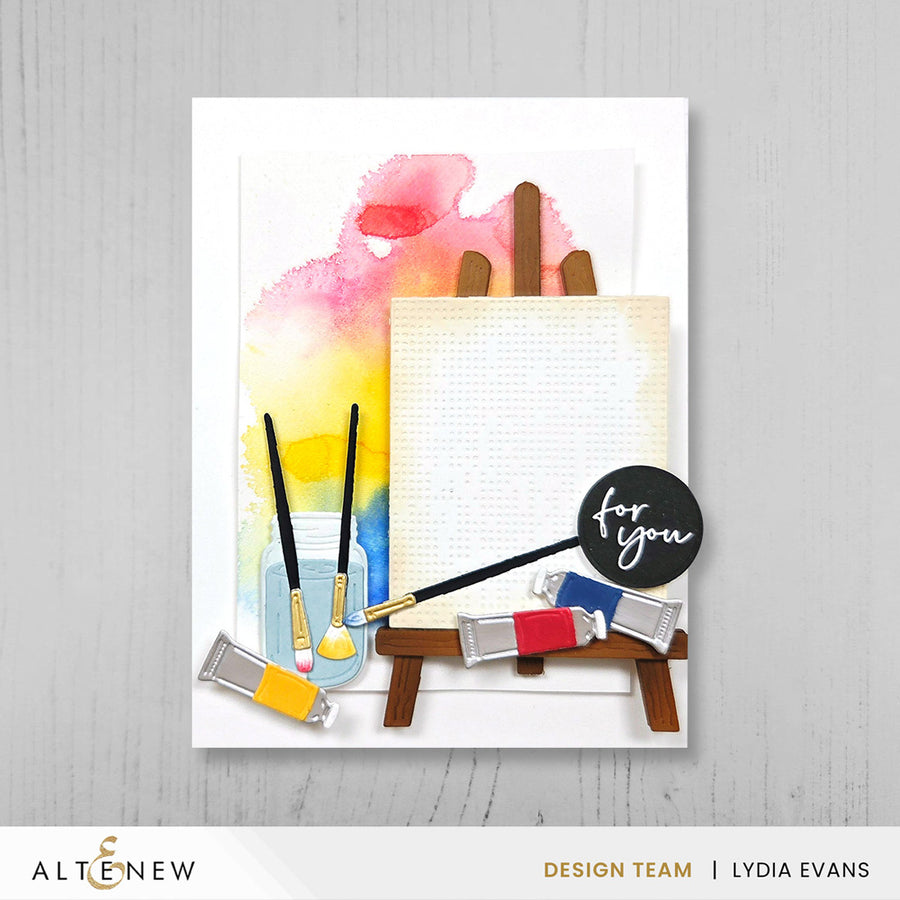 A handmade greeting card featuring a watercolor painting palette design with paintbrushes, paint tubes, a blank canvas on an easel, and a black circular badge that reads 'for you'. The card is displayed against a light gray wood-textured background, with the Altenew and Design Team Lydia Evans branding visible. Altenew One-Go: Solid Circular Sentiments Press Plate card ideas
