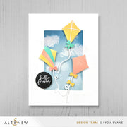A handmade greeting card featuring three colorful paper kites against a blue sky background with white clouds, displayed on a light gray wood-textured background. The card has a white border and a black circular sticker that reads 'hello friend'. Altenew One-Go: Solid Circular Sentiments Press Plate card ideas