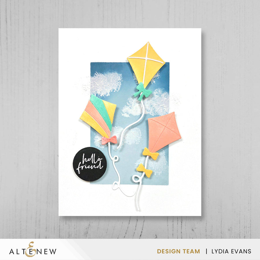 A handmade greeting card featuring three colorful paper kites against a blue sky background with white clouds, displayed on a light gray wood-textured background. The card has a white border and a black circular sticker that reads 'hello friend'. Altenew One-Go: Solid Circular Sentiments Press Plate card ideas